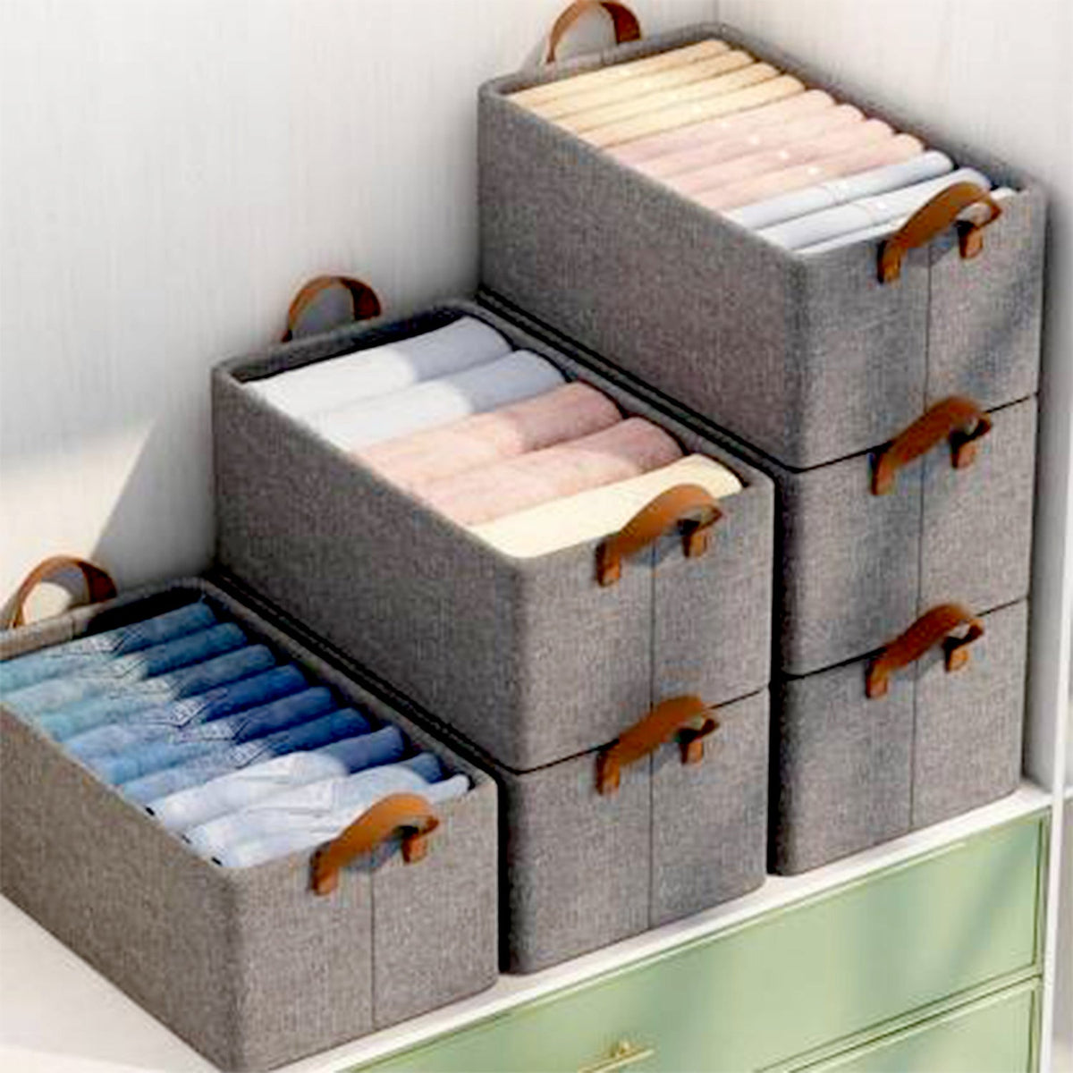 Three grey fabric storage boxes filled with folded clothes, stacked on a light green dresser.