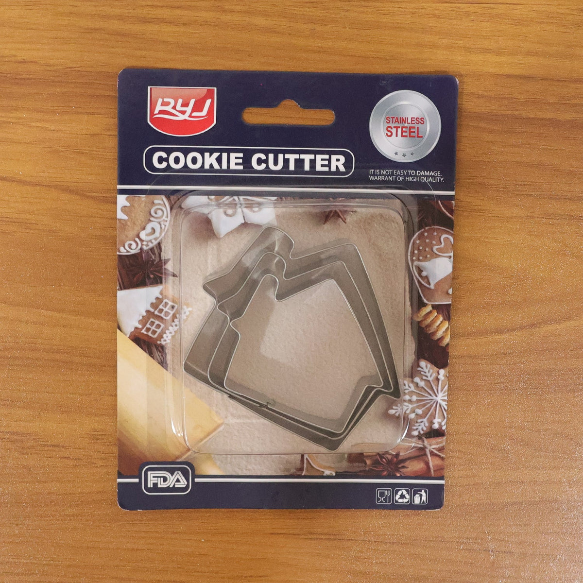 A packaged set of three stainless steel house-shaped cookie cutters on a light brown wooden surface.