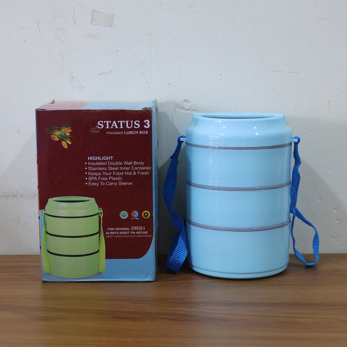 A light blue three-layer lunch box with a blue strap stands beside its red and blue packaging, featuring a green box image.