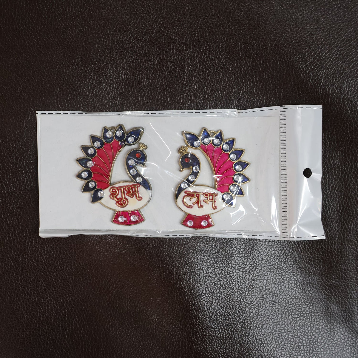 The pair of peacock Shubh Labh wall hangings are sealed in clear plastic packaging on a dark brown textured surface.