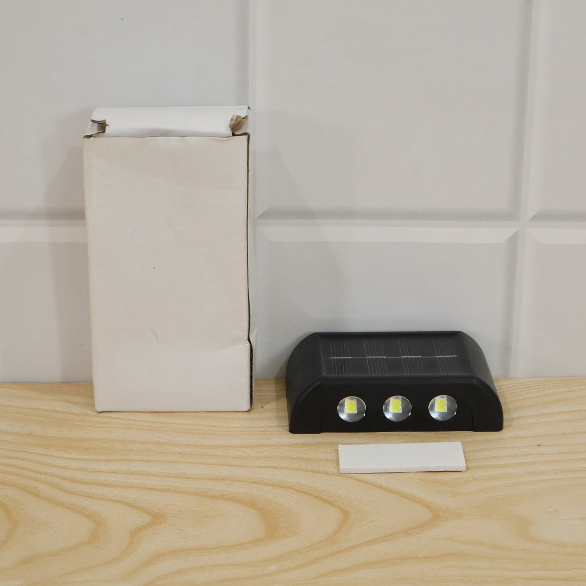 A black solar-powered LED wall light with a solar panel and three bright LEDs, a white adhesive strip, and a cardboard box