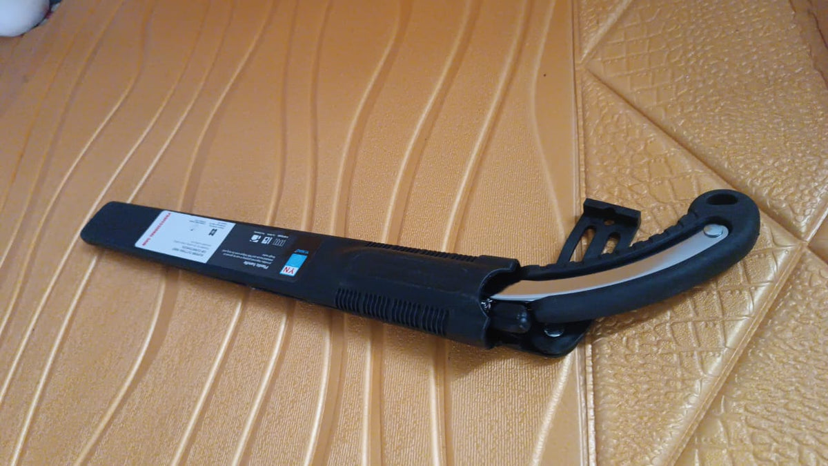 A black folding hand saw with a silver blade and its black cover are laid on a textured golden-brown surface.