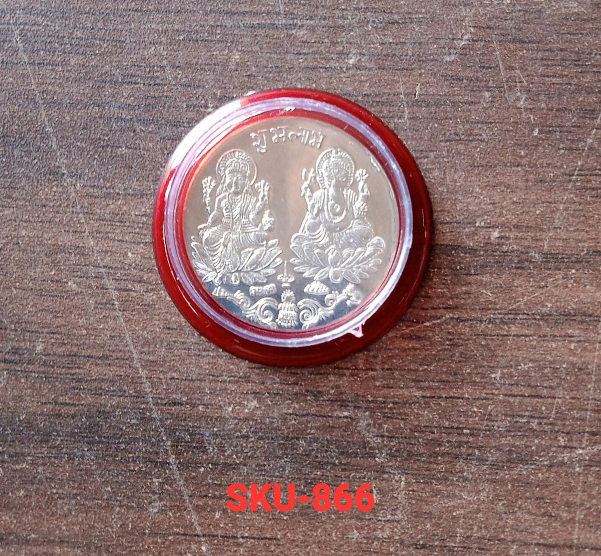 A silver coin with Lakshmi and Ganesha in a red and clear plastic case, resting on a wooden surface.