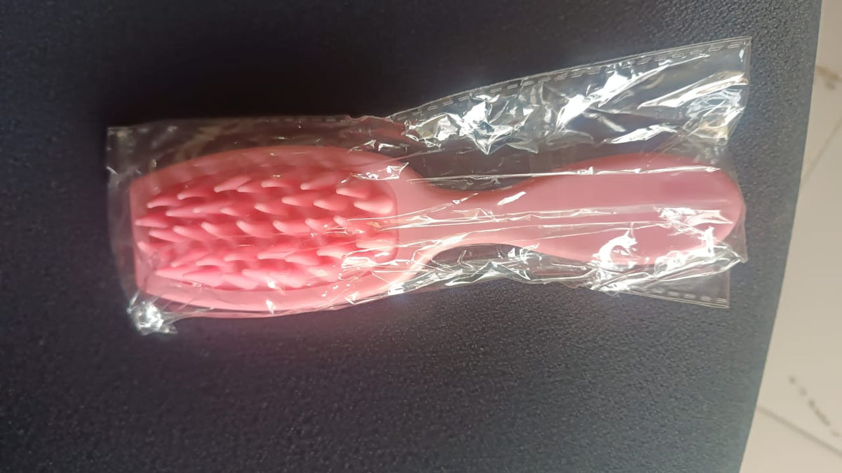 A pink silicone scalp massager brush is sealed in clear plastic packaging on a dark surface.