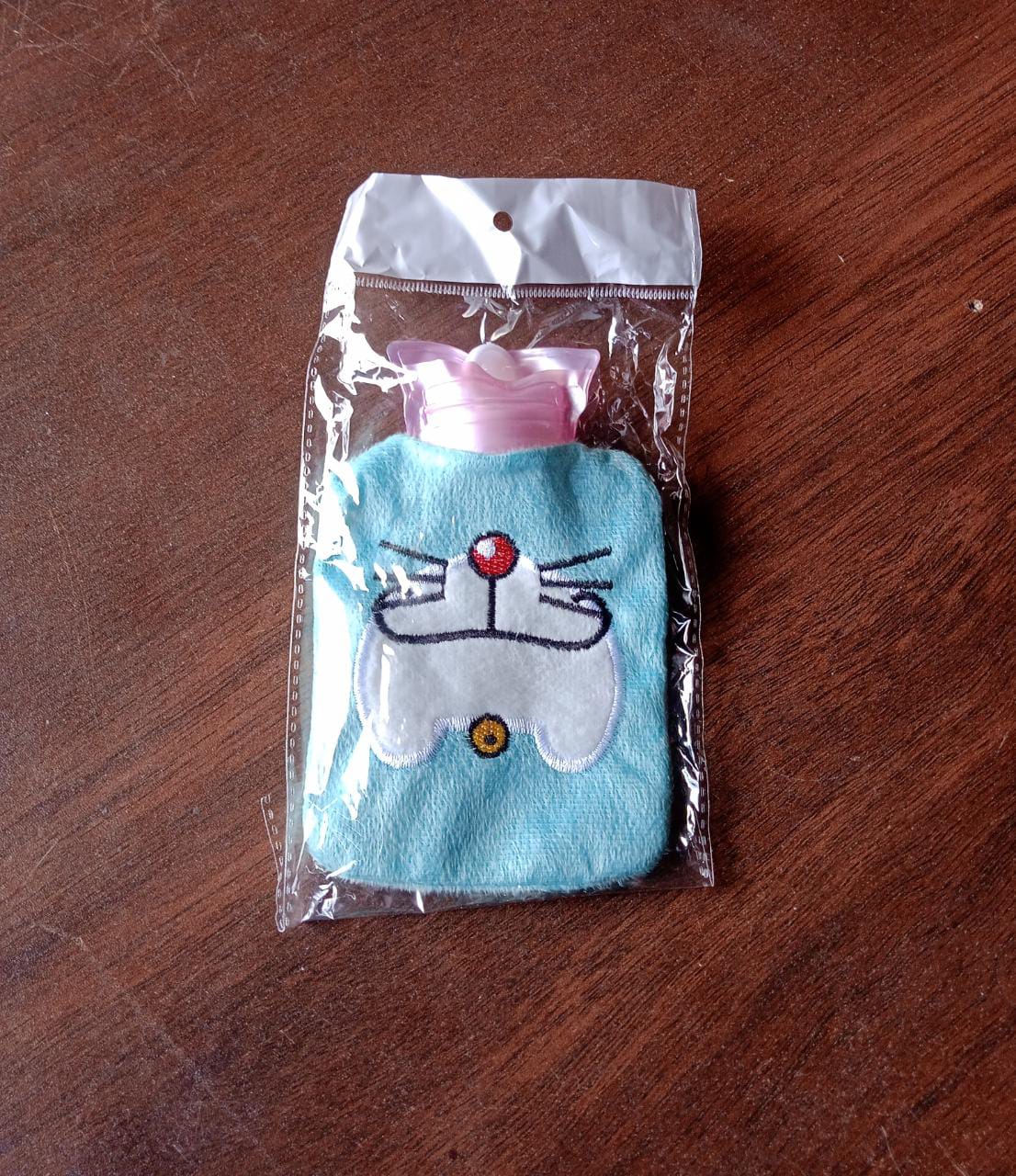 A light blue hot water bag with a Doraemon face design is sealed in clear plastic packaging on a wooden surface.