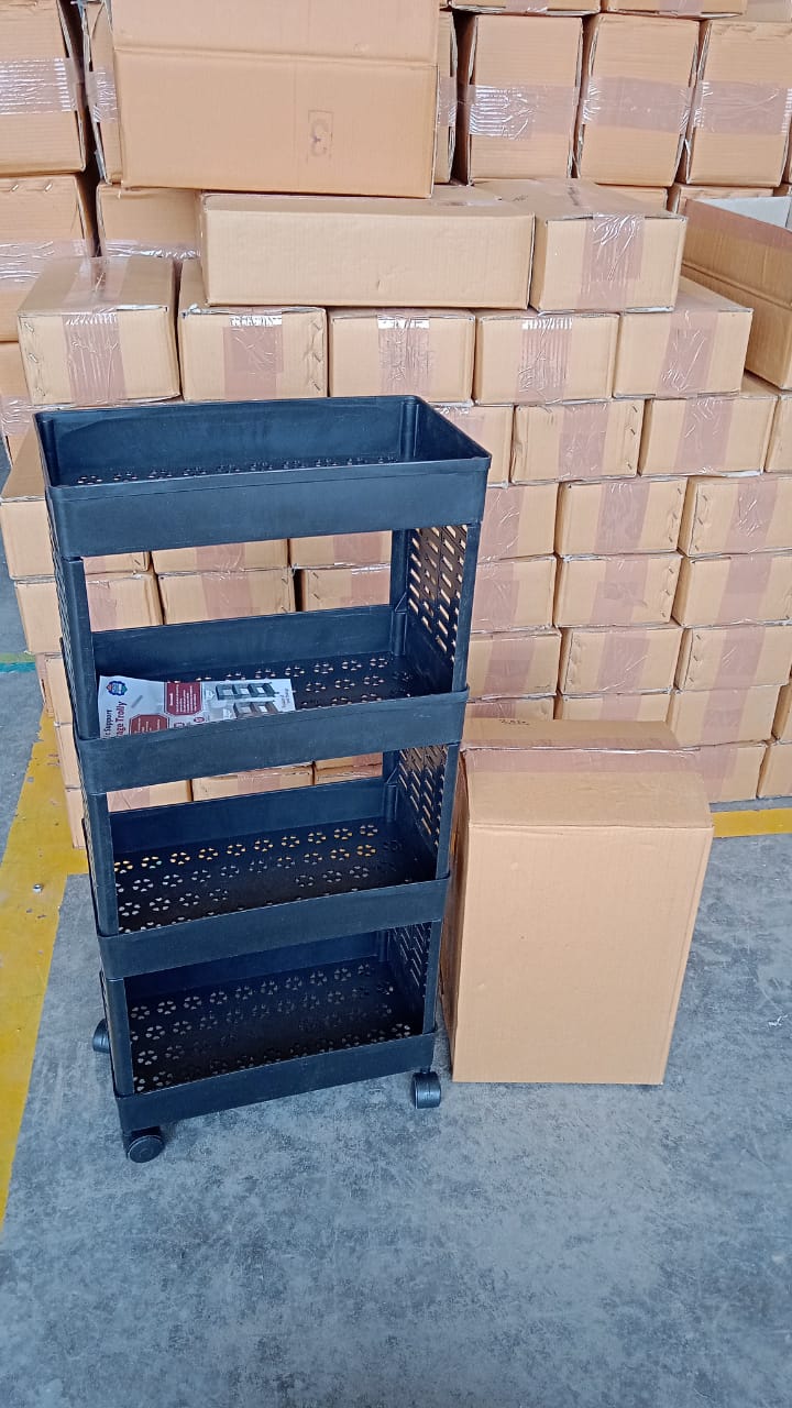 A black four-tier plastic utility trolley on wheels stands next to a brown box, surrounded by stacks of cardboard boxes in a