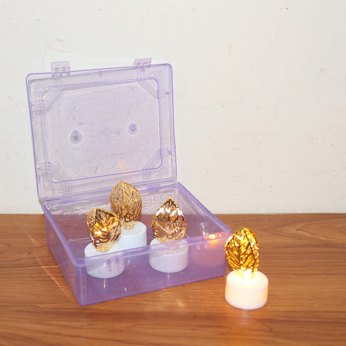 Three golden leaf LED candles are inside an open clear plastic box, with one candle outside on a wooden surface.