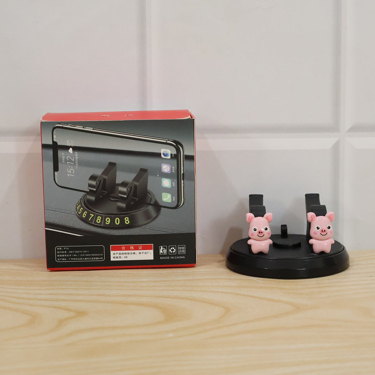 A black phone holder with two pink pig figurines next to its product box on a wooden surface.