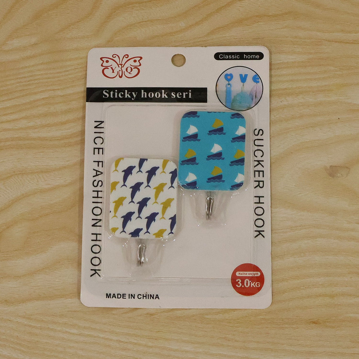 The packaged set of two adhesive hooks, featuring dolphin and sailboat designs, resting on a light wooden background.