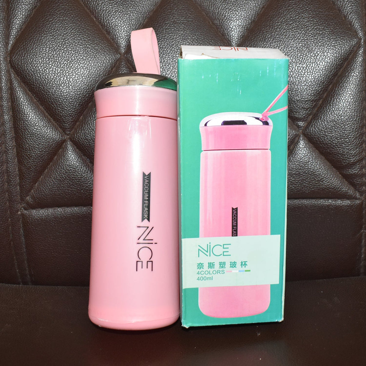 A pink vacuum flask with a silver lid and strap stands next to its retail box on a dark brown quilted surface.