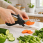 A person's hands use a black and white clever cutter to slice a carrot on a white countertop with vegetables around it.