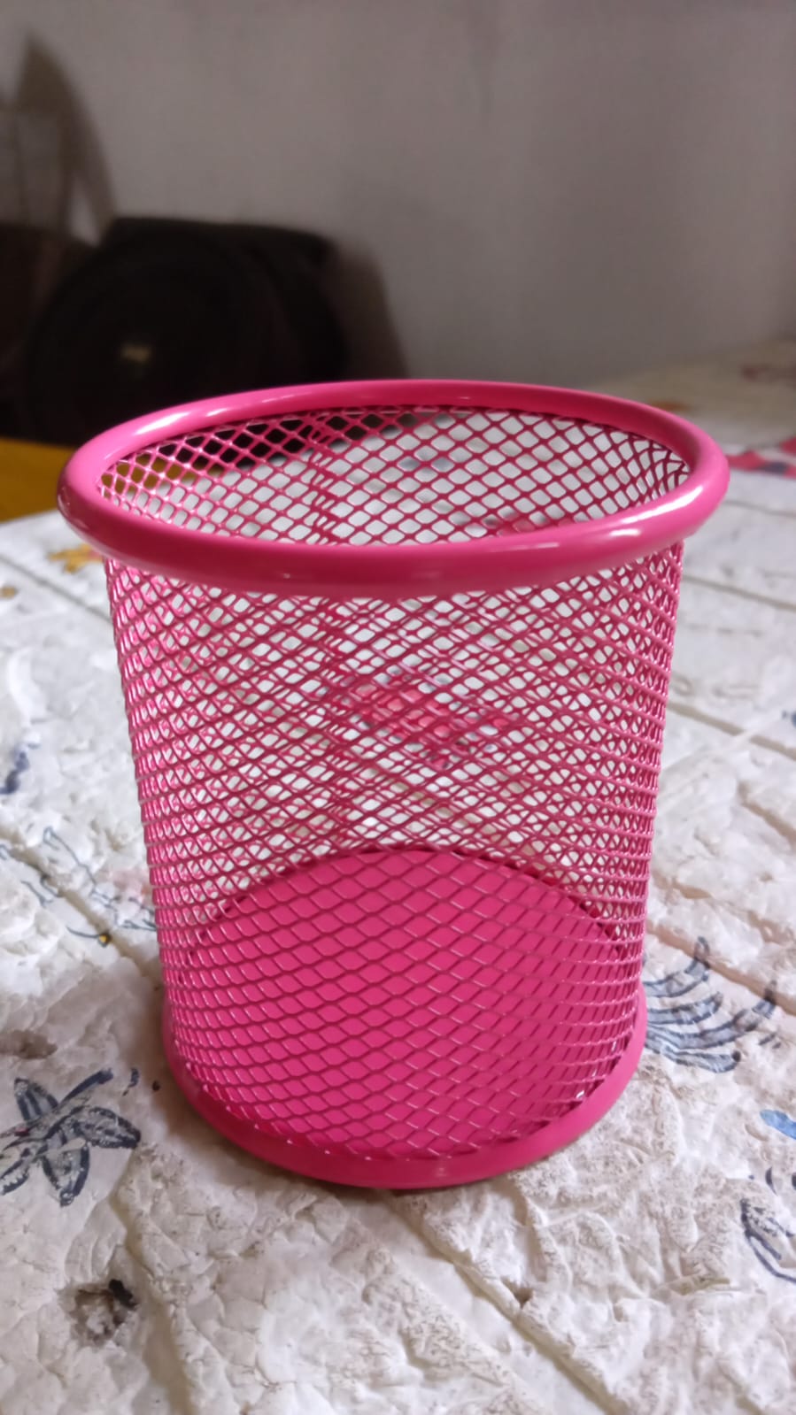A pink metal mesh pen holder rests on a patterned white and blue fabric surface.