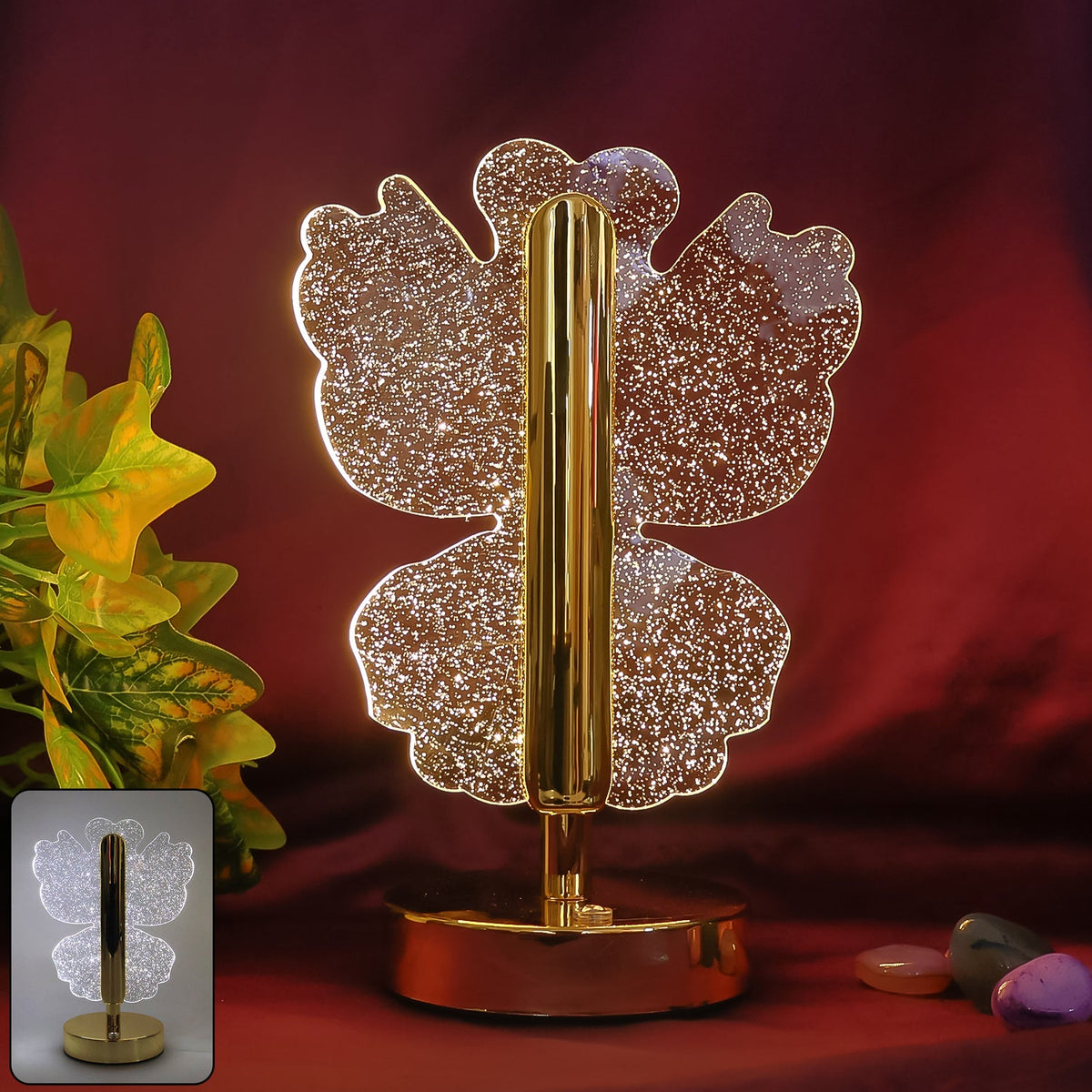 A glowing crystal butterfly lamp with a gold stem and base, against a dark red background with green foliage and small rocks.