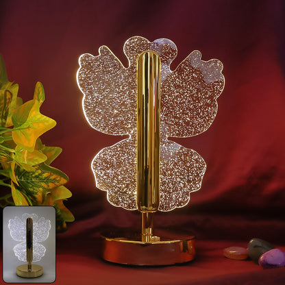 A glowing crystal butterfly lamp with a gold stem and base, against a dark red background with green foliage and small rocks.