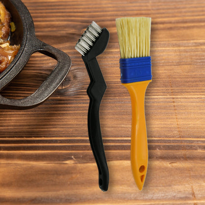 A black grill cleaning brush and a yellow and blue basting brush stand side-by-side on a rustic wooden surface.
