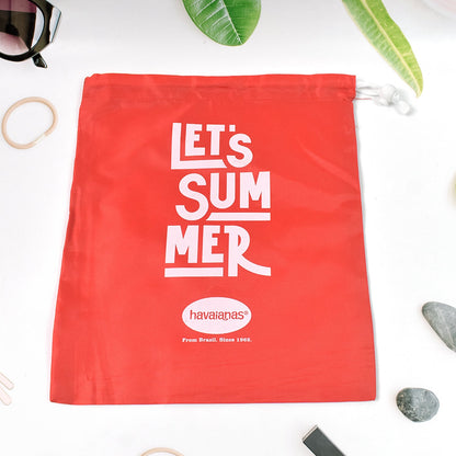 A red drawstring bag with "LET'S SUMMER" and Havaianas logo, on a white surface with green leaves and small grey stones