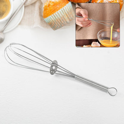 A silver spring coil whisk is displayed on a white surface with a blurred background of baked goods and person whisking