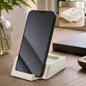 A black smartphone stands upright in a cream plastic desktop holder on a wooden table, with an inset showing the holder.
