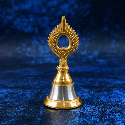 A small brass and steel pooja bell with a peacock feather design stands against a textured dark blue background