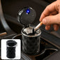A hand places a lit cigarette into a black car cup holder ashtray with a blue LED light.
