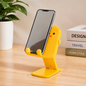A yellow cartoon duck phone stand holds a smartphone on a wooden desk next to a plant and books.