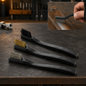 Three mini wire brushes with black handles and different bristle types on a dark, textured surface, with one brush cleaning