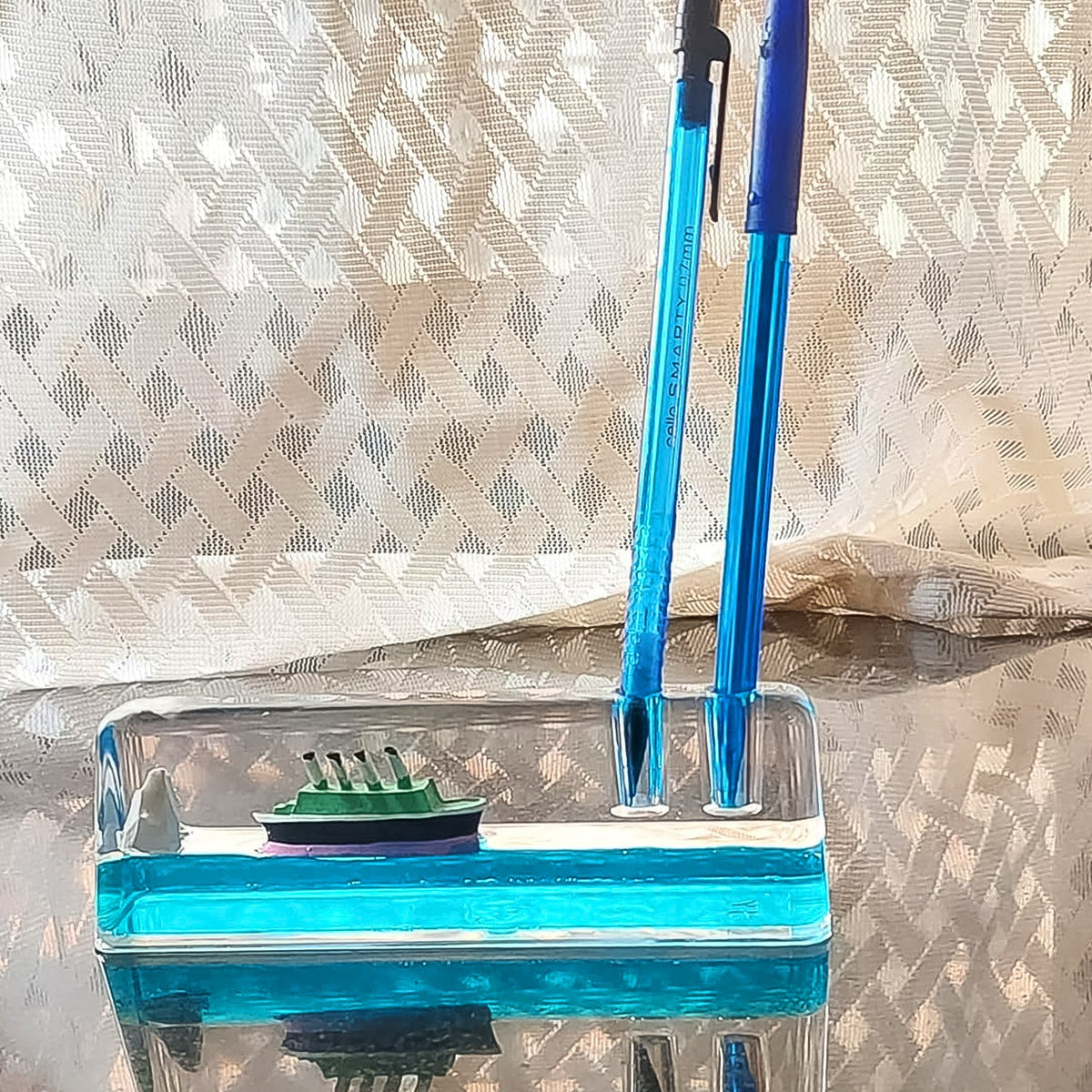 A clear liquid motion paperweight with a ship and iceberg, also serving as a pen holder with two blue pens inserted.