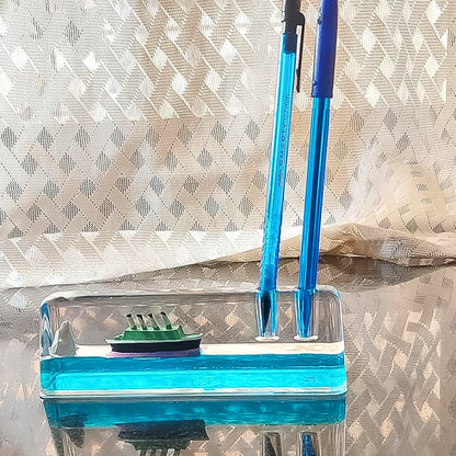 A clear liquid motion paperweight with a ship and iceberg, also serving as a pen holder with two blue pens inserted.