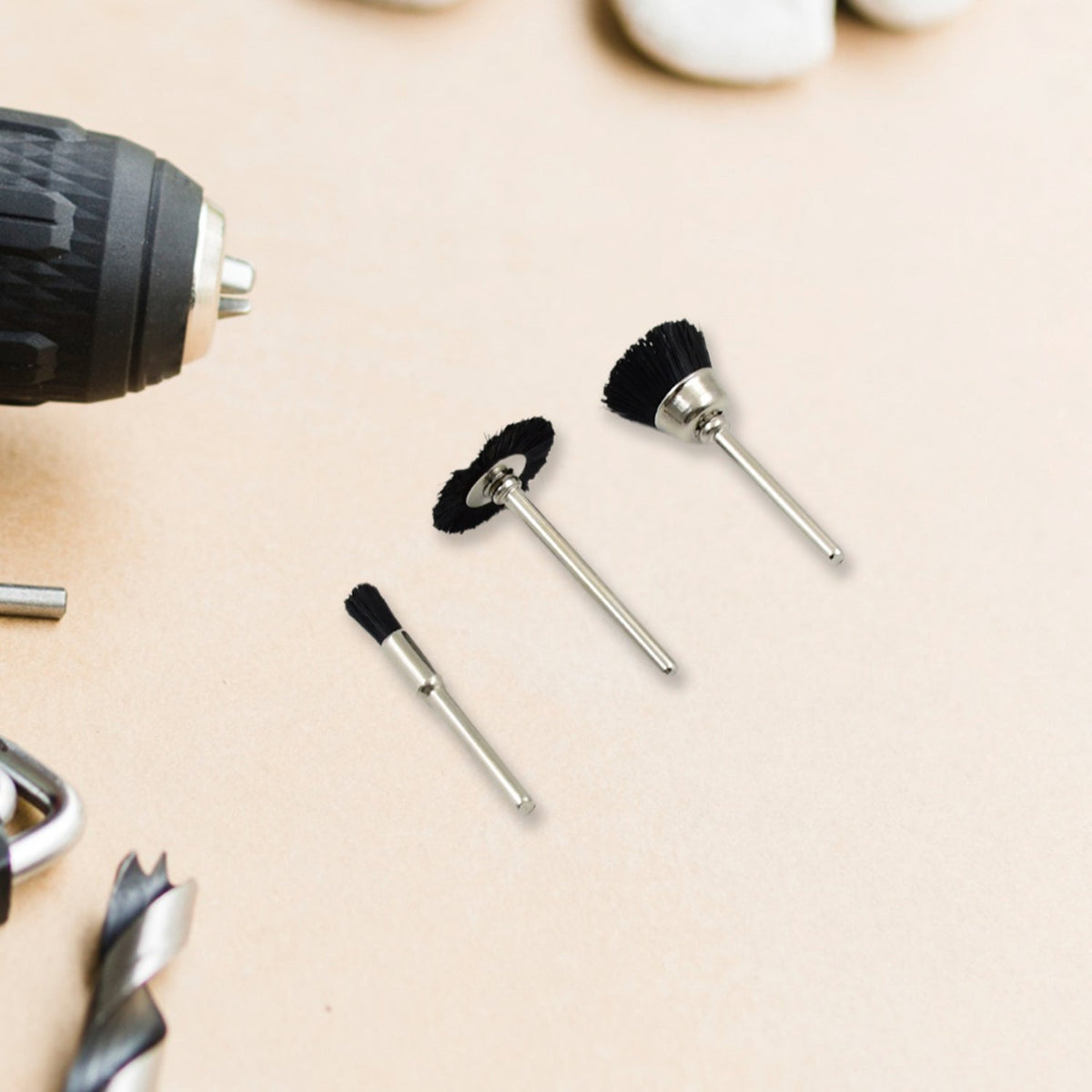 Three black nylon brushes with silver shafts are arranged on a light brown surface next to a drill and other tools.