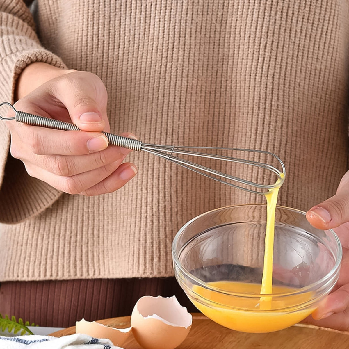A person in a beige sweater holds a small silver whisk, dripping beaten egg into a clear glass bowl.