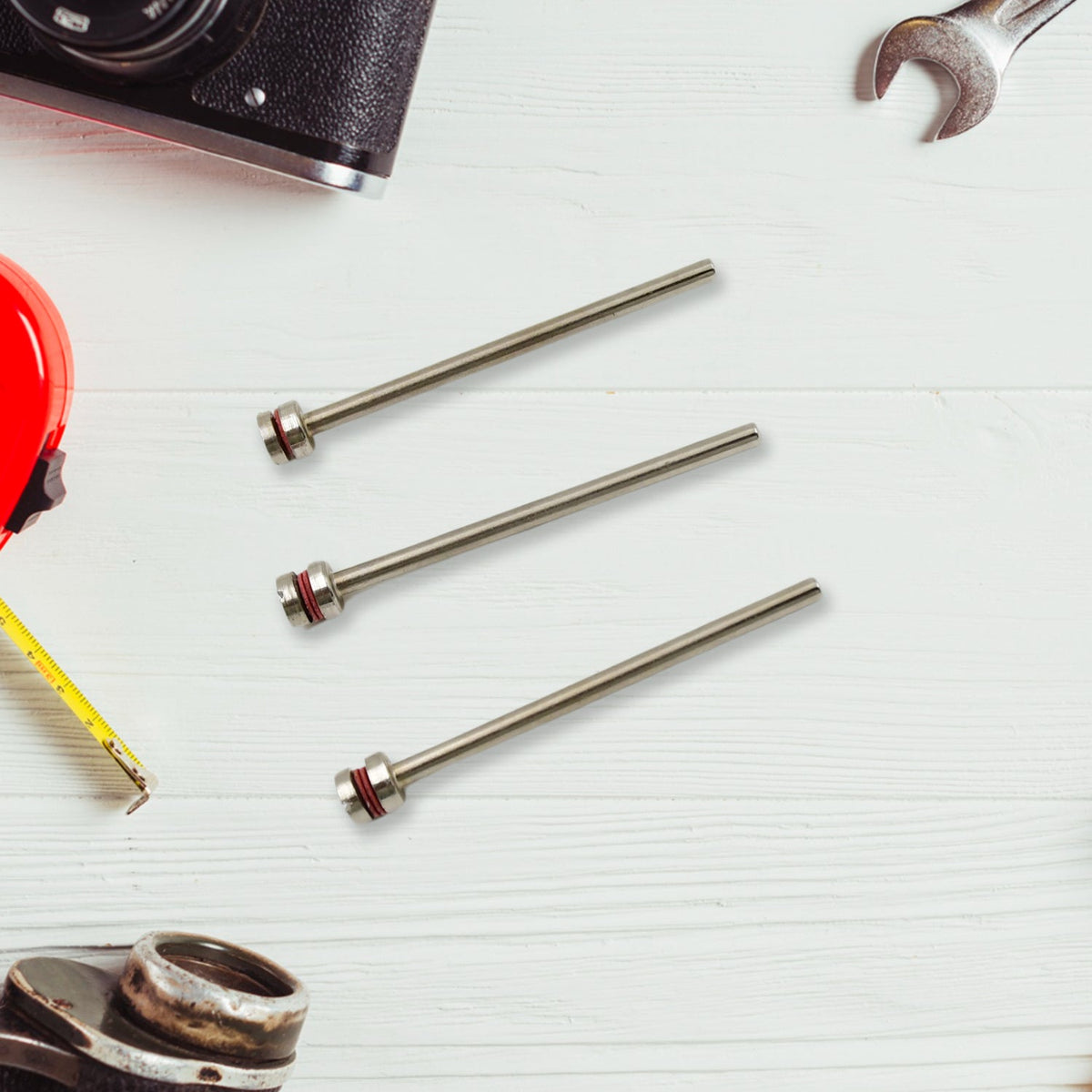 Three silver mandrels with red bands arranged on a white wooden surface, surrounded by a camera, tape measure, and wrench.