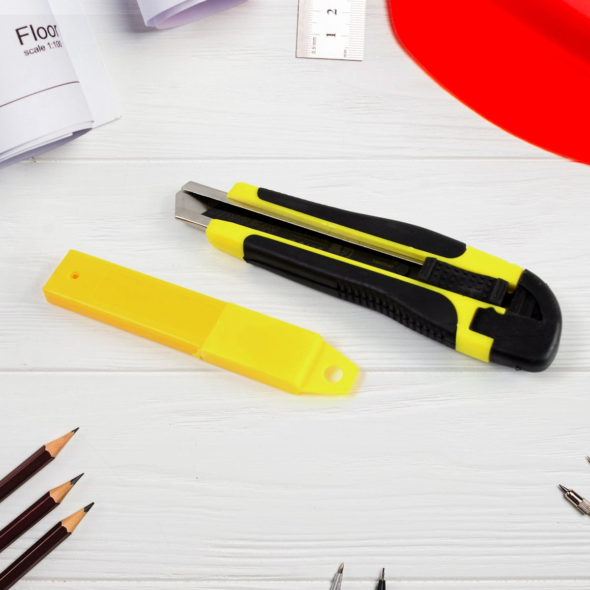A yellow and black utility knife with a yellow blade cover on a white wooden surface with pencils.