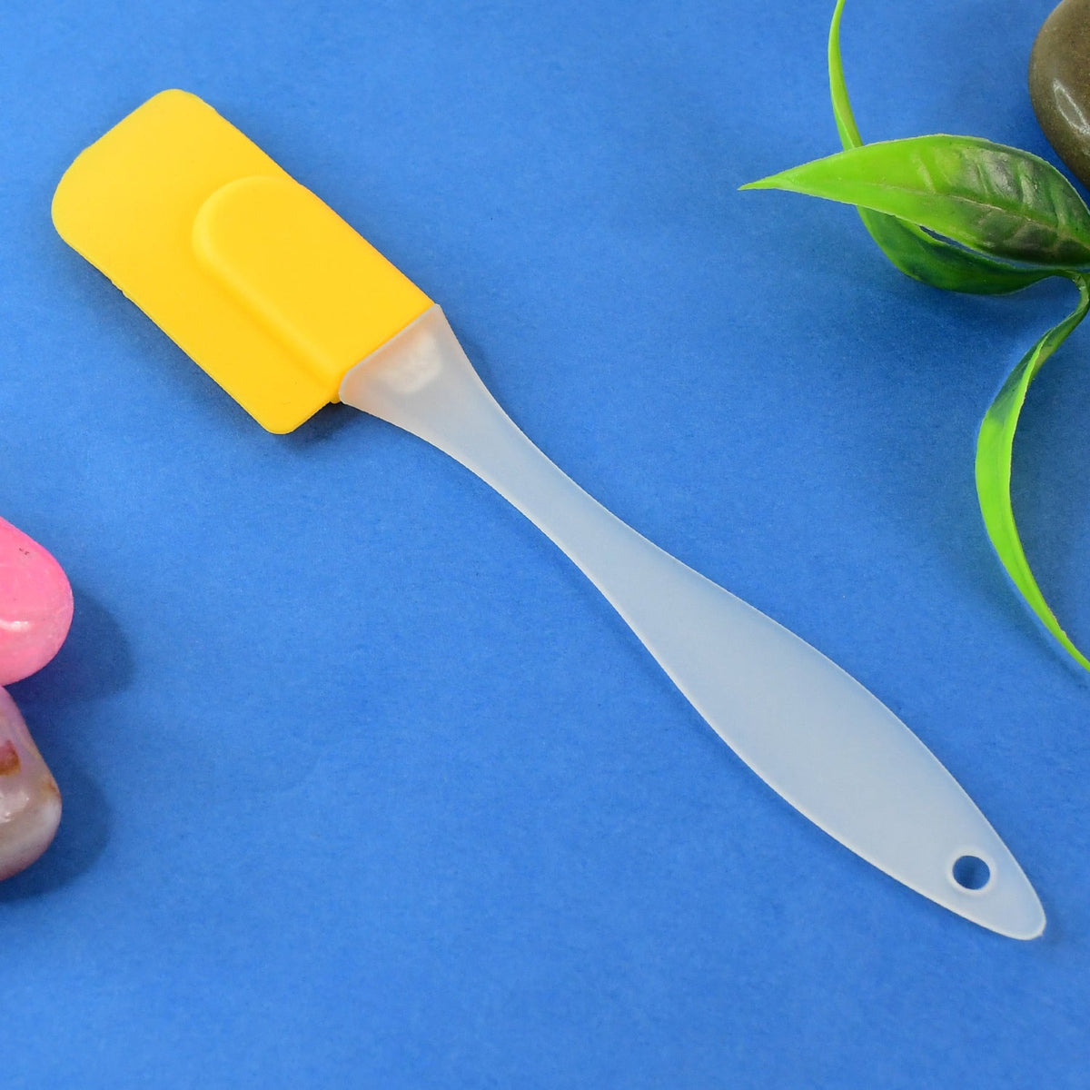 A yellow silicone spatula with a translucent handle lies on a vibrant blue surface, with green leaves and a pink object.