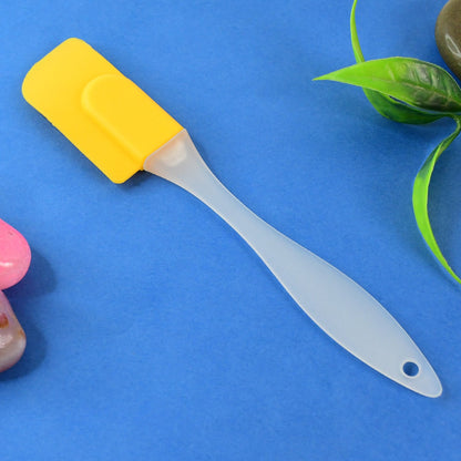 A yellow silicone spatula with a translucent handle lies on a vibrant blue surface, with green leaves and a pink object.