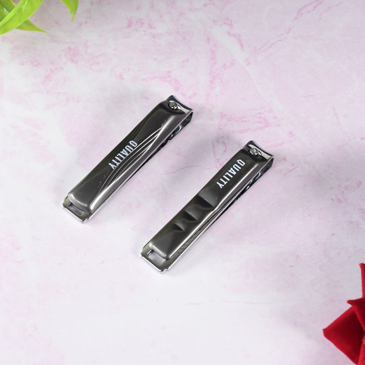 Two closed black and silver nail clippers with "QUALITY" on the handles, on a pink marble surface.