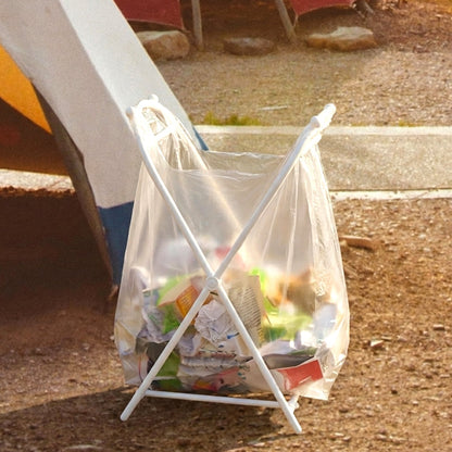 White plastic garbage bag holder with a clear bag full of garbage, outdoors on a dirt path.