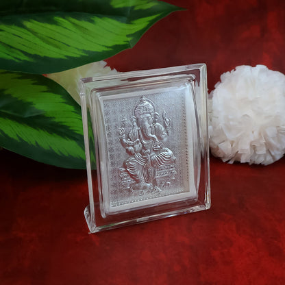 Silver-plated Ganesha idol in an acrylic frame, with a white fluffy decoration and green leaves on a red surface.
