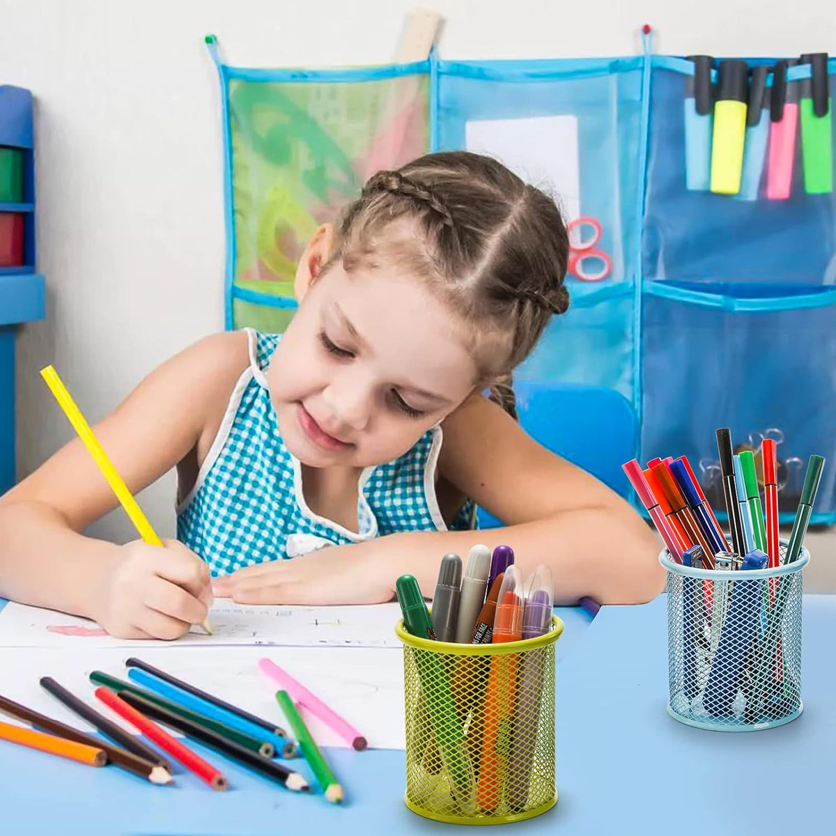 A young girl draws at a blue table with green and light blue mesh pen holders filled with markers.