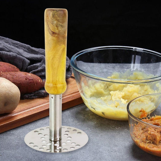 A stainless steel masher with a light wooden handle stands beside bowls of mashed potatoes and sweet potatoes.