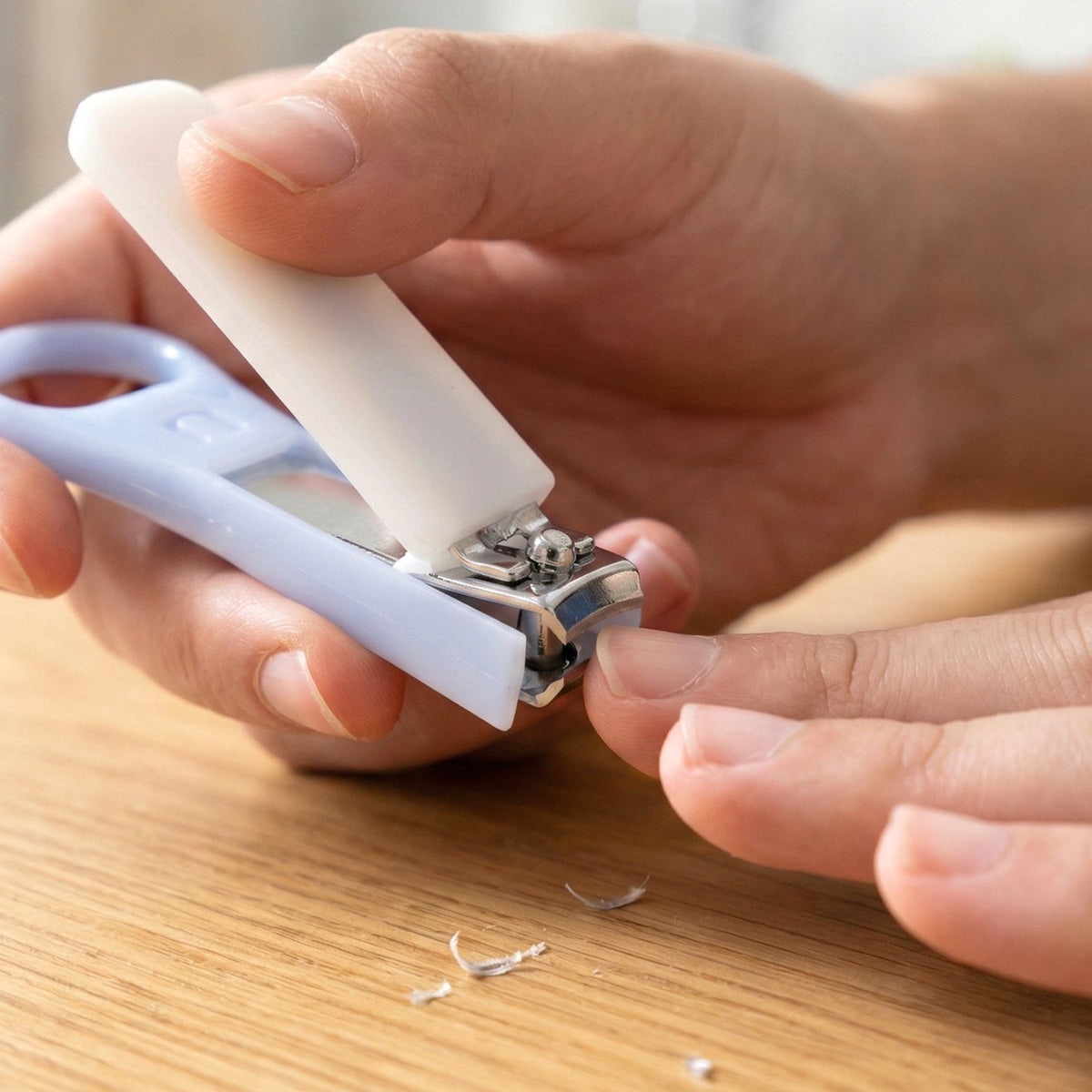 A person's hands using the light blue and white nail clipper to trim a fingernail, with nail clippings on the table.