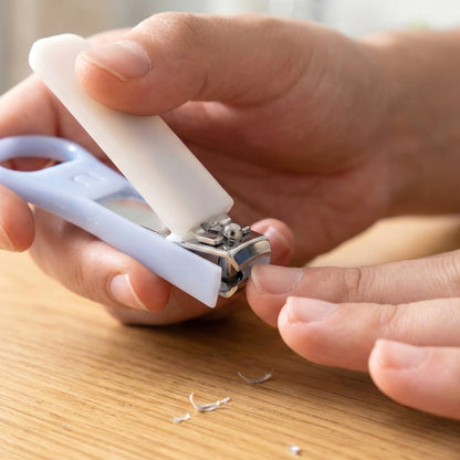 A person's hands using the light blue and white nail clipper to trim a fingernail, with nail clippings on the table.