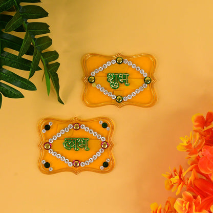 Two yellow Shubh Labh wall hangings with green text and jewels, on a yellow wall with green and orange foliage.