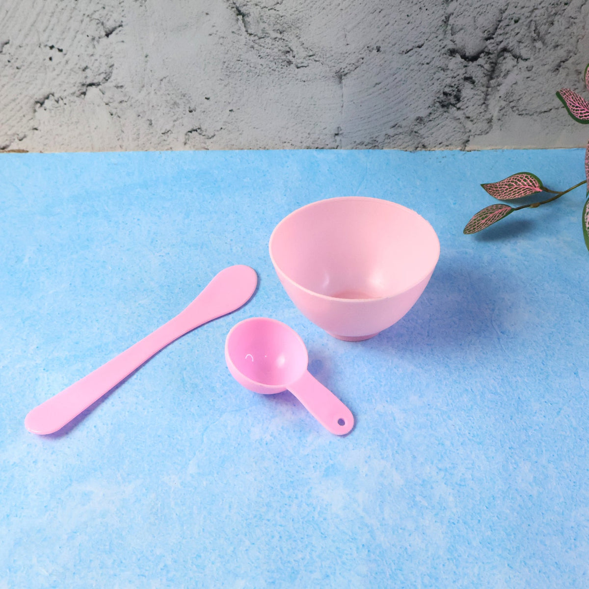A pink mixing bowl, spatula, and measuring spoon are displayed on a textured blue surface.