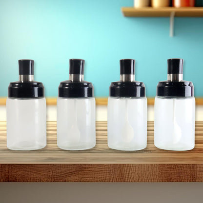 Four transparent spice jars with black lids and white spoons on a wooden shelf with a blue wall.