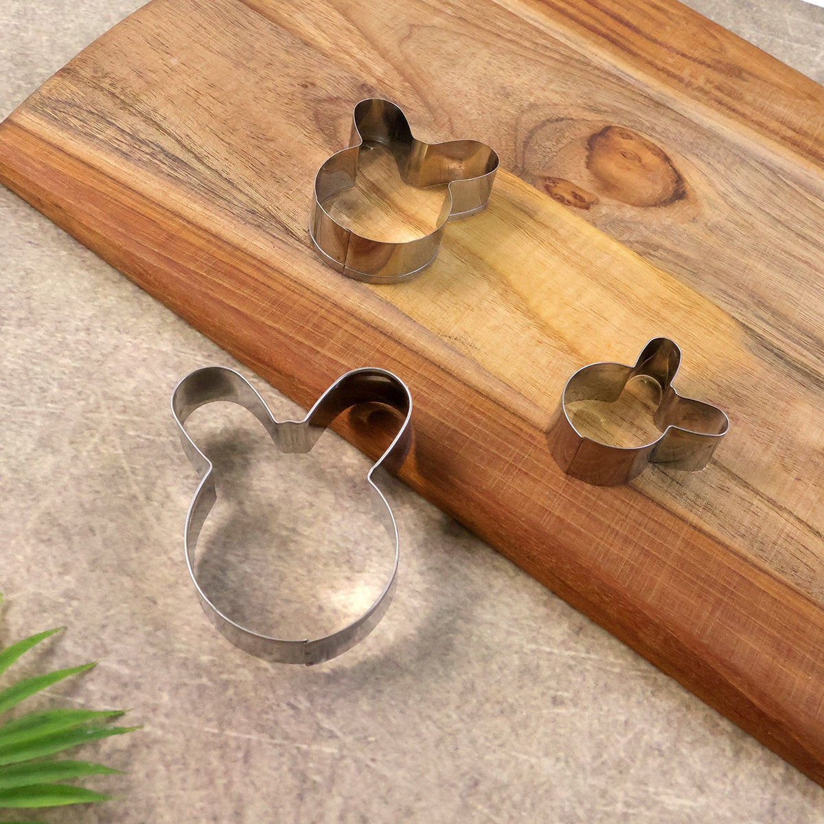 Three bunny-shaped stainless steel cookie cutters in different sizes arranged on a wooden cutting board with a green leaf