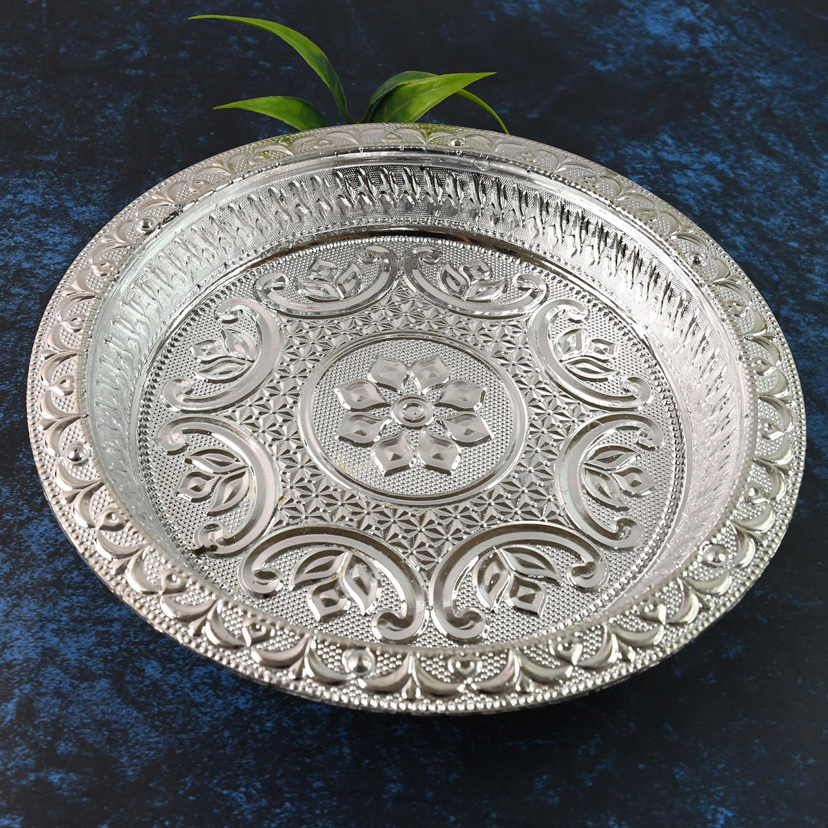 A round silver-colored plastic pooja thali with intricate embossed floral and geometric patterns, with green leaves in the