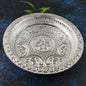 A round silver-colored plastic pooja thali with intricate embossed floral and geometric patterns, with green leaves in the