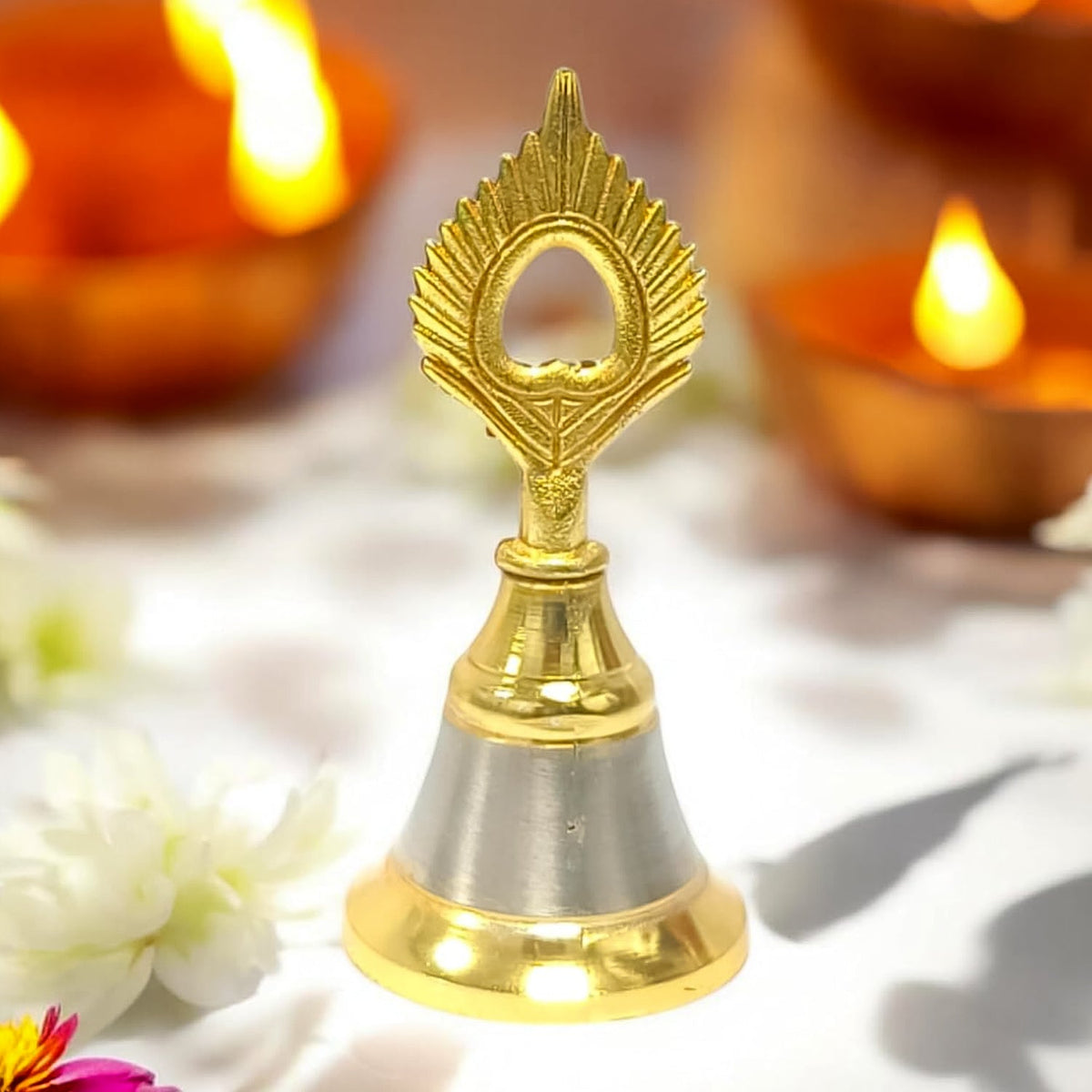 A small brass and steel pooja bell with a peacock feather design stands on a white surface surrounded by lit diyas and white