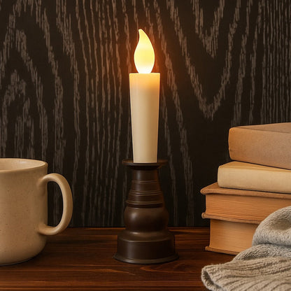A cream LED candle with a flickering flame sits in a dark bronze holder next to a mug and books.