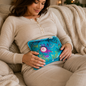 A woman in a beige sweater holds a teal floral electric hot water bag on her stomach while sitting on a couch.